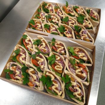 Two trays of rolled sandwich wraps filled with herbs, tomato and red onion, arranged in neat rows.